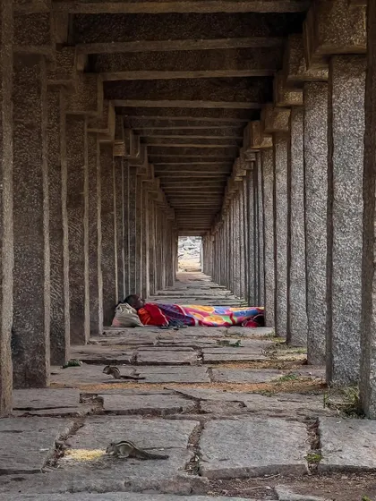 A person sleeps in the long, pillared corridor of a temple, sharing the space with squirrels. This candid shot captures a quiet moment of daily life persisting within the historic ruins.
