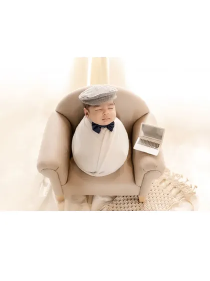 The little boss. A newborn, swaddled and wearing a dapper cap and bow tie, sleeps in a miniature armchair with a tiny laptop. This humorous and adorable themed shot shows the creative possibilities of a newborn session.