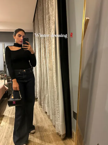 A winter dressing mirror selfie in a black cutout top and dark jeans.