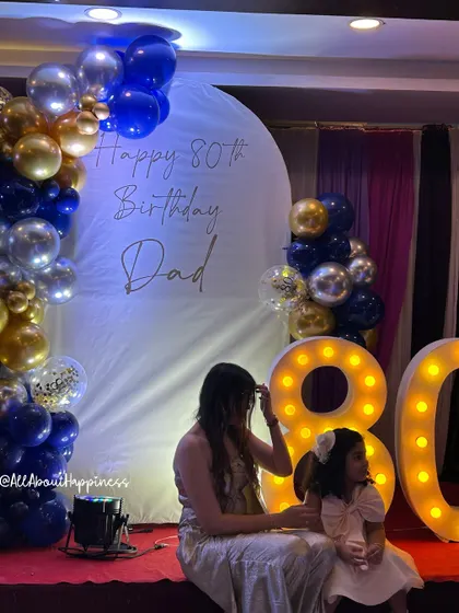 An elegant setup for an 80th birthday celebration, featuring a white arch backdrop with custom calligraphy, a blue, gold, and silver balloon garland, and large marquee numbers.