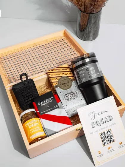 A modern and stylish groomsman hamper in a cane-lidded wooden box. This one is packed with tech and treats, including a JBL speaker, luxury socks, gourmet coffee, and a "Groom Squad" card.