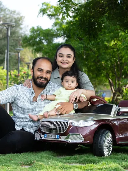 An outdoor family portrait with parents and their baby posing with a miniature toy car in a green park.
