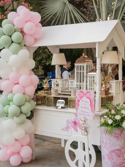 A charming white cart used as a dessert or candy station, decorated with a pastel balloon garland.