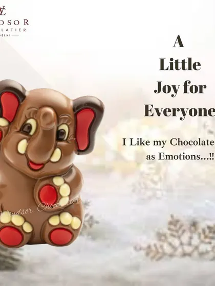 A joyful, waving baby elephant sculpture, crafted from milk chocolate with colorful red accents on its ears and feet.
