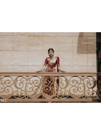 A wide, cinematic portrait of the bride on an ornate balcony, looking like royalty in her traditional red and gold saree.