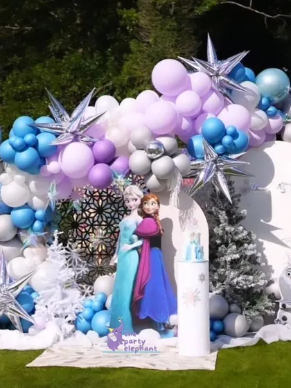 I created a spectacular winter wonderland for this Frozen-themed birthday. The backdrop features character cutouts, snow-covered trees, and a massive balloon garland in shades of blue, purple, and silver with starburst accents.