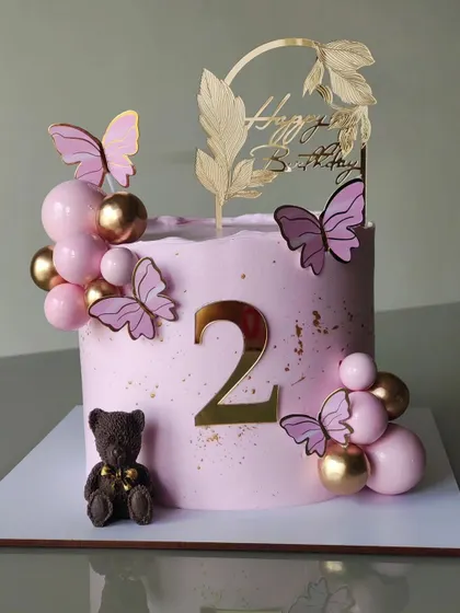 A sweet and simple butterfly-themed cake for a second birthday. The soft pink buttercream is decorated with butterflies, a chocolate bear, and a cascade of pink and gold spheres.