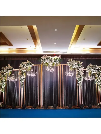 A glamorous reception decor with tall golden frames, dark curtains, and elegant chandeliers. The white floral arrangements on top add a touch of softness to the modern structure.