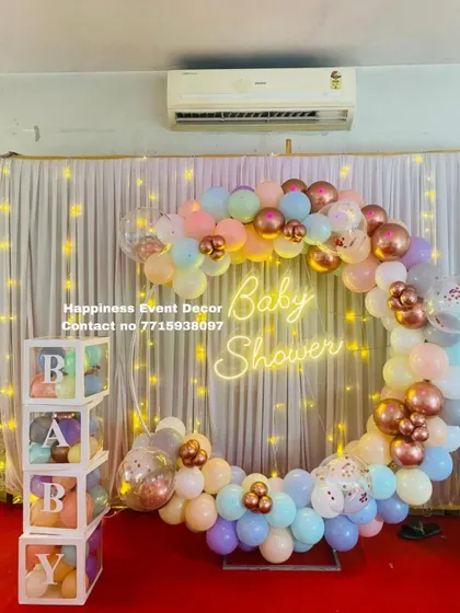 This baby shower decor uses a full balloon ring with pastel and rose gold chrome balloons. The setup includes a 'Baby Shower' neon sign and transparent 'BABY' boxes filled with balloons.