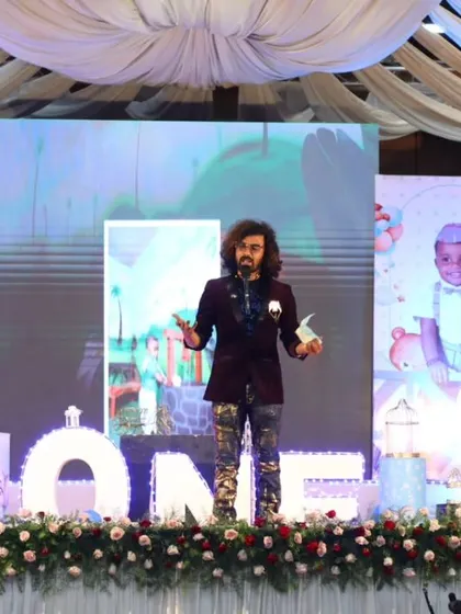 Addressing the audience at a first birthday party, with a beautifully decorated stage as the backdrop.