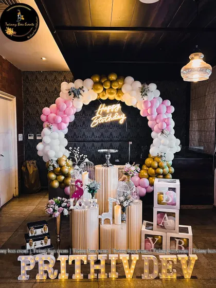 A full view of Prithivdev's first birthday setup. It features a circular balloon arch in pink, white, and gold, fluted pedestals, "BABY" blocks, and his name in large marquee letters.