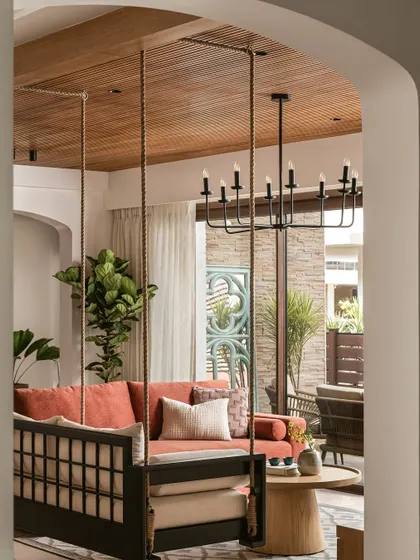 Seen through an archway, the informal living room showcases a mix of comfortable seating, including a rust-colored sofa and a modern swing. The wood-paneled ceiling adds warmth and texture to the space.