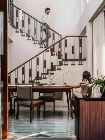 **The Glow House, Ahmedabad:** The double-height dining area is defined by the staircase, which acts as a sculptural element. The railing's design creates a pattern of light and shadow, adding a layer of visual interest to the daily routine of moving through the home.