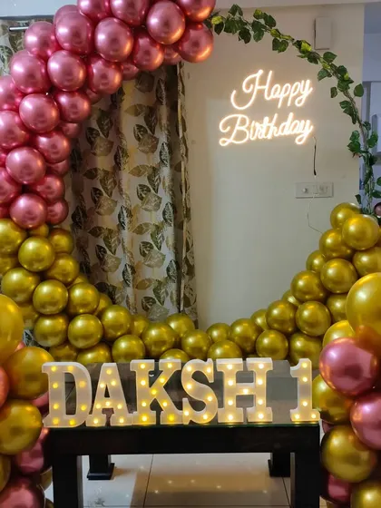 A metallic pink and gold balloon arch with a neon sign, set up for a first birthday at home.