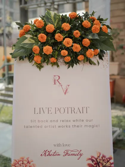 A repeat of the "Live Portrait" sign, topped with a fresh arrangement of marigolds, seamlessly integrating this unique activity into the overall Haldi decor.