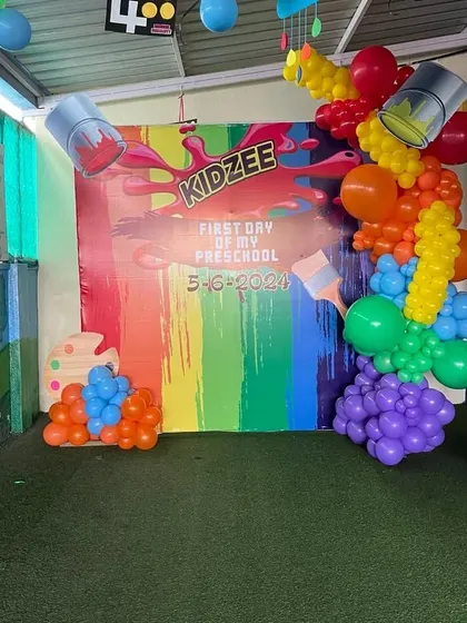 An "Art Splash" themed backdrop for a Kidzee preschool event. The decor features a colorful printed backdrop and a creative balloon installation designed to look like paint spilling from buckets.