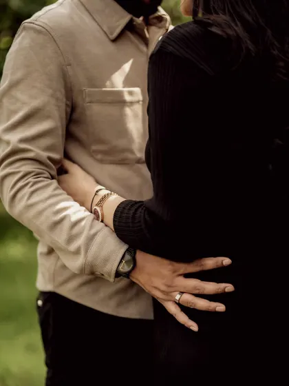 A close-up in warm monochrome, focusing on the couple's embrace and their hands on the baby bump. The sunlight filtering through the trees adds a beautiful, gentle light.