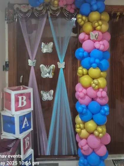 A door decoration for a baby celebration, featuring a vibrant balloon garland in pink, blue, and yellow, with butterfly accents and 'BABY' blocks.