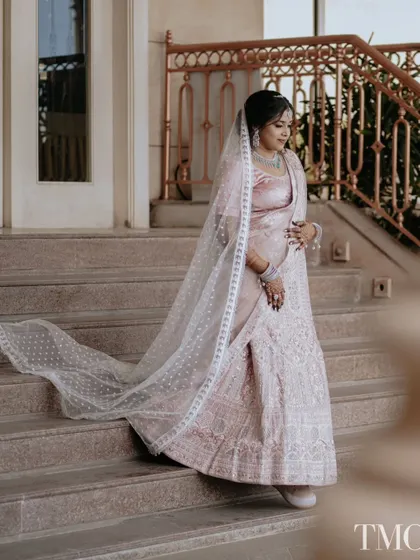 A princess moment on the stairs. The bride descends a grand staircase, her pastel lehenga and long veil trailing behind her, creating a classic and graceful bridal portrait.