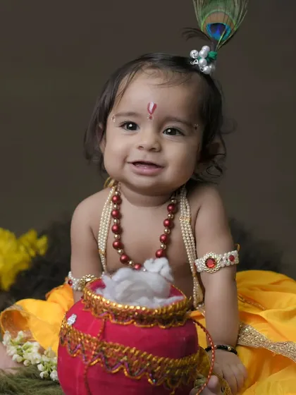 A sweet and serene portrait of a baby dressed as Krishna. The simple, dark background helps the colorful costume and adorable expression stand out.