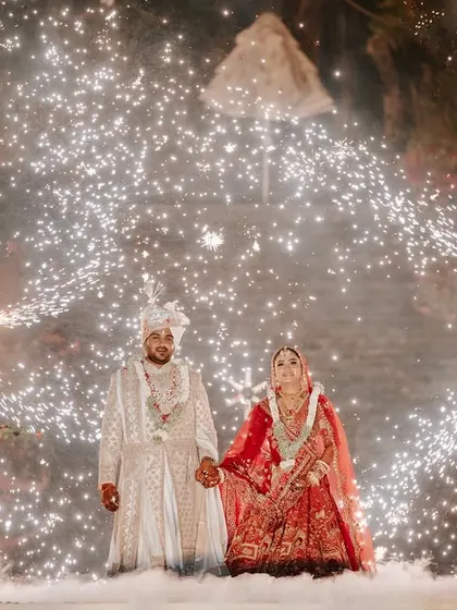 The couple stands together amidst a shower of sparks and ethereal low-lying fog. This wide shot showcases the full scale of the pyrotechnic display we designed for their Varmala.