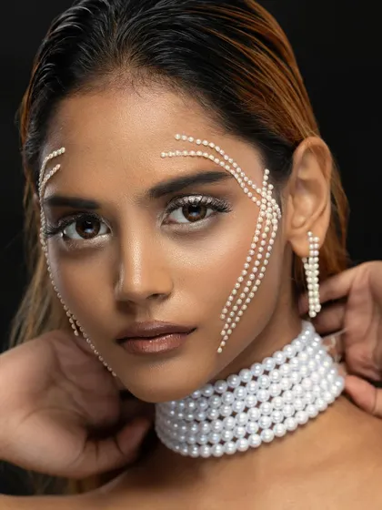 An editorial beauty shot focusing on intricate details. The pearl face adornments and choker create a look of avant-garde elegance against a clean, dark background.