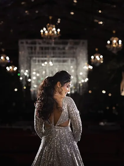 A stunning back-shot of the bride in her shimmering sangeet lehenga, looking out over the reception hall. The chandeliers create a beautiful bokeh, highlighting her elegant silhouette.