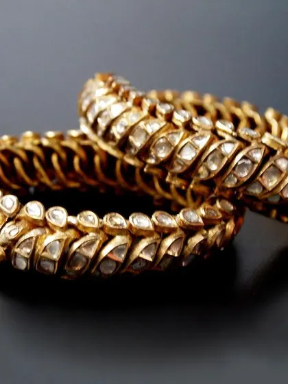 The pair of royal 'Shrankhla Jodi' bangles, presented against a dark surface to emphasize the warm glow of the antique gold and the sparkle of the diamonds.