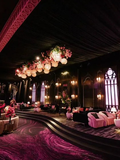 A dramatic lounge area with a dark, moody atmosphere. The space features plush pink sofas on a tiered platform, gothic-style arched windows, and a ceiling adorned with hanging flowers and lanterns.