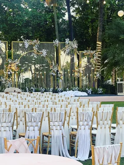 An elegant daytime reception setup with a mirrored gold backdrop. The design reflects the surrounding palm trees and sky, creating a unique and glamorous effect for an outdoor event.