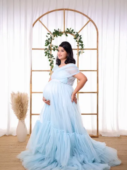 A full-length portrait of a mother-to-be in a light blue gown, framed by our studio's arched window prop for a classic and elegant composition.