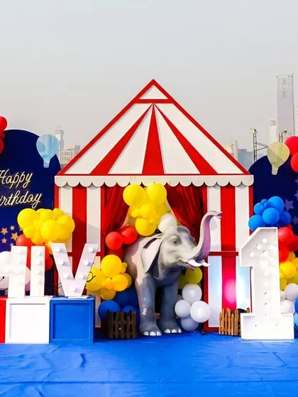 A grand circus-themed birthday setup against a city skyline, complete with a big top tent, animal props, and marquee letters spelling out the birthday boy's name.