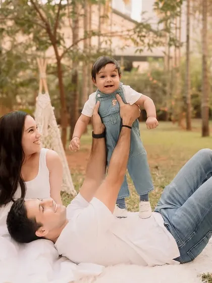 A father holds his son up in the air while mom watches, all enjoying a beautiful day in the park.
