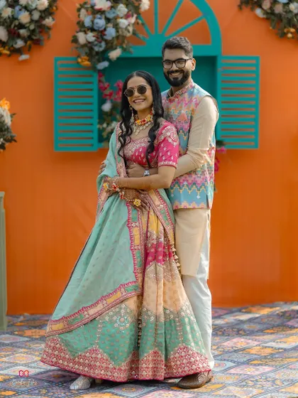 A full-length portrait of the couple in their vibrant Mehendi outfits, standing before a colorful, festive backdrop.