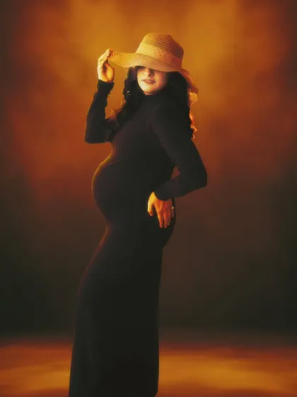 The glow of motherhood, framed in light. This portrait is a masterpiece capturing the warmth, strength, and beauty of an expecting mother in a simple black dress and hat.