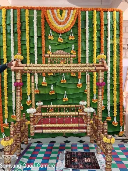 A traditional naming ceremony setup with a backdrop made of fresh banana leaves and marigold garlands. The wooden cradle is decorated to match, creating a vibrant and auspicious setting.
