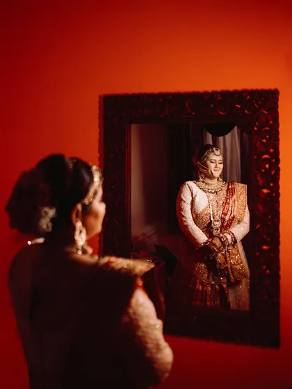 A beautiful mirror portrait of the bride. Her reflection is perfectly captured, showcasing her full bridal attire and serene smile. The red background adds a touch of passion and drama.
