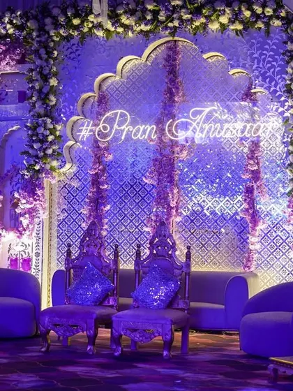 A spectacular stage setup for a Sangeet or reception night. The backdrop features intricate silver jali work, illuminated by purple light, with a custom neon hashtag for the couple.