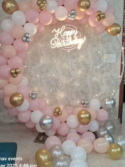 A beautiful birthday backdrop with a circular frame and a balloon garland in pastel pink, white, and gold. The setup is illuminated by a 'Happy Birthday' neon sign and fairy lights.