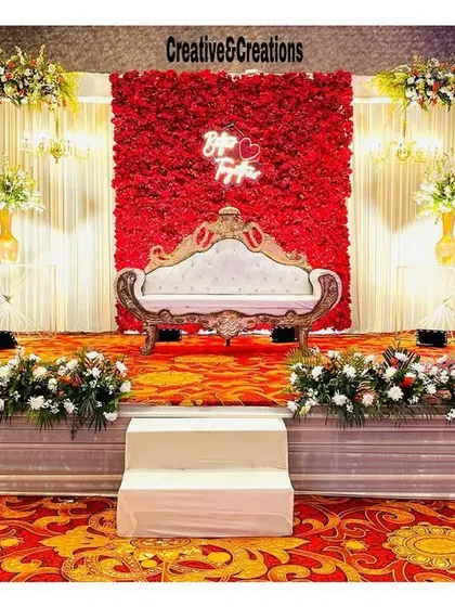 A beautiful stage design combining a red rose wall with a white curtain backdrop. The ornate sofa and floral arrangements at the base complete this classic wedding look.