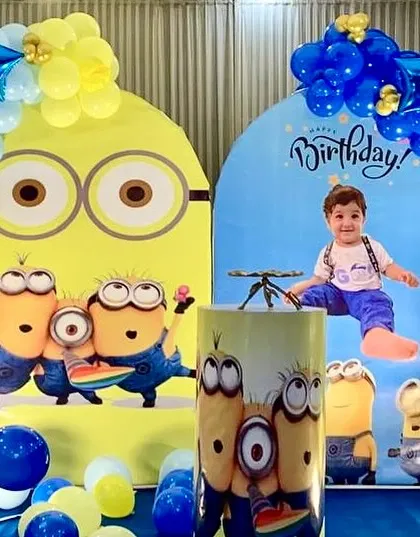 A fun-filled Minions-themed birthday party. The decor includes large backdrops with Minion characters, blue and yellow balloon arrangements, and a themed pedestal.