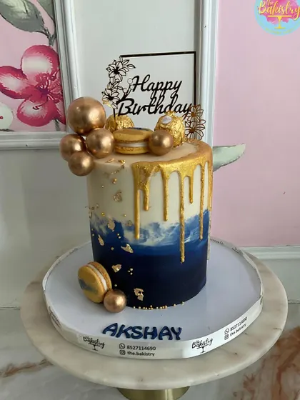 A stylish blue and white cake with a gold drip, topped with golden macarons and Ferrero Rocher for Akshay.