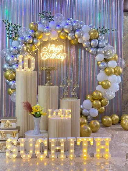 A 10th birthday party with a full circular balloon arch in silver and gold. The setup includes multiple plinths, marquee letters, and a neon sign against a shimmer curtain.
