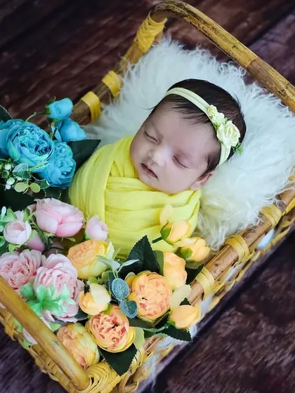 A baby in a yellow swaddle, nestled in a rectangular basket filled with colorful flowers.