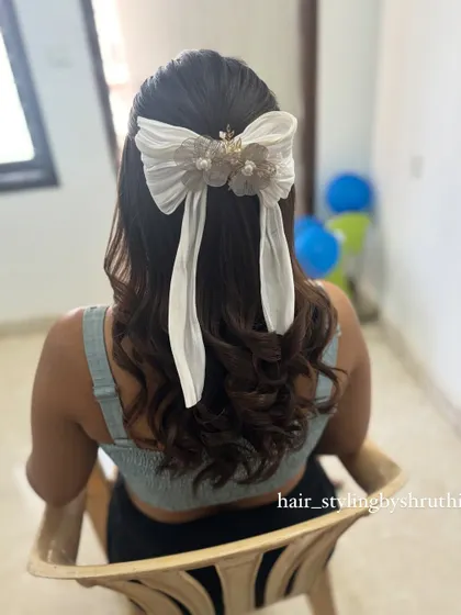 A trendy half-up style featuring loose waves and a large, pleated white bow accessory. This look is modern and chic, perfect for a fashion-forward client.