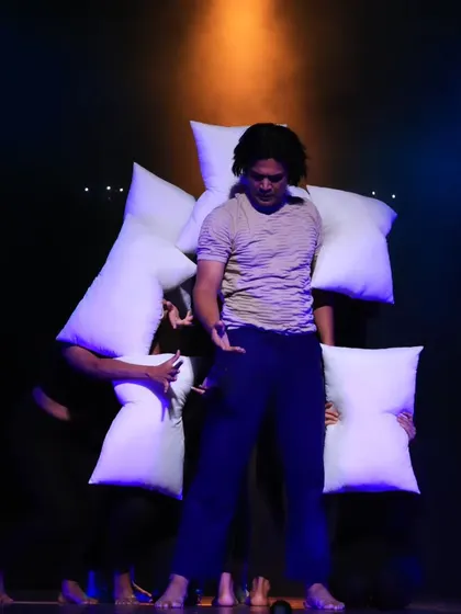 A performer from "ME & MY PILLOW" is surrounded by other dancers holding pillows. The orange and black background creates a striking visual contrast in this contemporary piece.