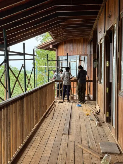 Workers install window frames on the upper-level balcony. The extensive use of wood for the floors, walls, and railings creates a warm and cohesive architectural language throughout the project.