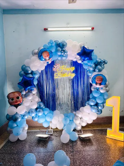 A Boss Baby themed first birthday decoration. The setup includes a circular balloon arch in blue and white, a shimmer curtain, and Boss Baby character foil balloons.