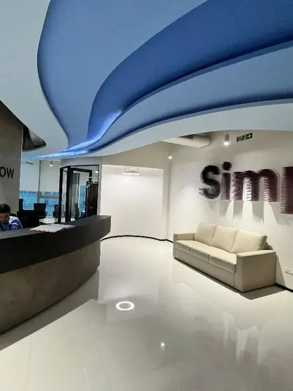 The reception area for Kissflow features a concrete-finish desk and a dynamic, wave-like ceiling element. The wall graphic "Simple" reinforces the company's core product philosophy in a bold, visual way.