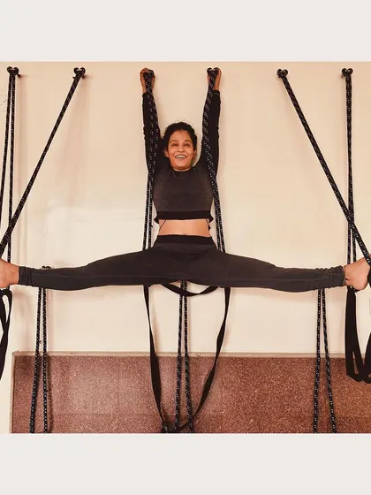 A student practicing Samakonasana (Center Splits) with the support of a rope wall. The ropes help to hold the body's weight, allowing for a gradual and safe opening of the hips and groin.